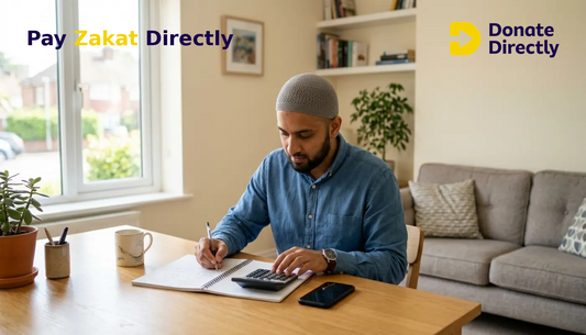 Muslim man in UK home calculating Zakat with calculator and phone at a bright table