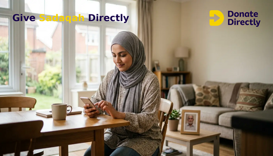 UK Muslim woman giving sadaqah on her phone at home with a framed photo of a loved one nearby