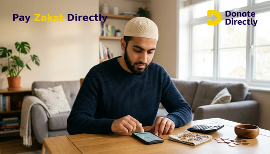 British Muslim man calculating Zakat on smartphone at home with coins and jewellery on table